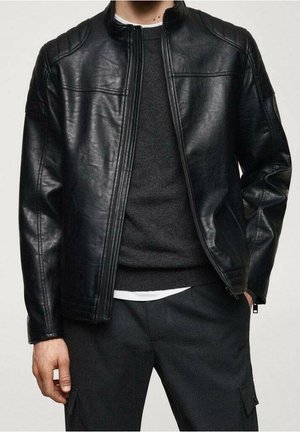 Homme portant une veste en cuir noire par-dessus un pull noir et un pantalon sombre, debout avec une main dans la poche et une main détendue sur le côté.