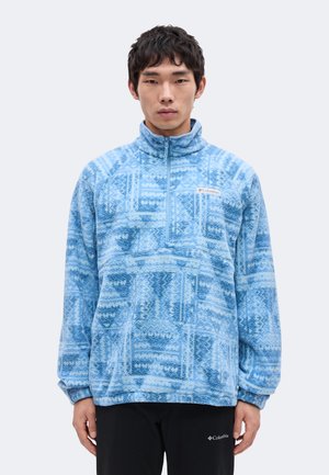 Muž oblečený v modrom vzorovanom Columbia vlnenom pulóvri so zipsom do polovice a čiernych nohaviciach Columbia, stojaci pred jednoduchým svetlým pozadím.
