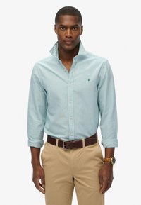 Camisa de botones a rayas azul claro con cuello, que presenta un pequeño logo verde en el pecho, combinada con pantalones beige y un cinturón marrón.