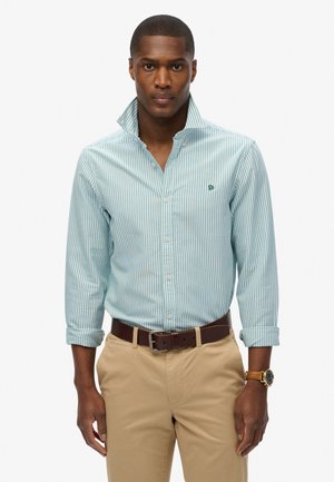 Light blue striped button-up shirt with a collar, featuring a small green logo on the chest, paired with beige pants and a brown belt.