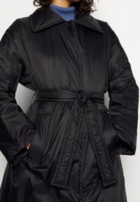 Černý péřový kabát s opaskem v pase, velkým límcem, dvěma bočními kapsami a hladkým, lesklým povrchem.