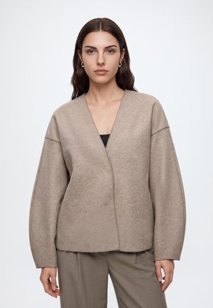 Donna con lunghi capelli castani che indossa orecchini a cerchio dorati e una giacca beige larga sopra pantaloni taupe, in piedi davanti a uno sfondo chiaro e semplice.