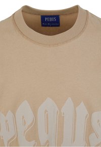 Beige színű kerek nyakú pulóver, texturált felszínt és feltűnő fehér feliratot tartalmaz. A címke "PEQUS" feliratú, standard bordázott gallérral rendelkezik.
