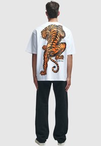 Hvit oversized t-skjorte med et stort oransje og svart tigertrykk på ryggen, kombinert med mørke joggebukser og hvite joggesko.