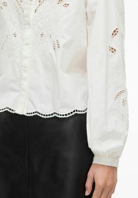 Balta kokvilnas blūze ar ziedu izšuvumu un izgriezuma detaļām, viļņotu apakšmalu un garām pūkainām piedurknēm. Kombinēta ar melnu ādas svārku.