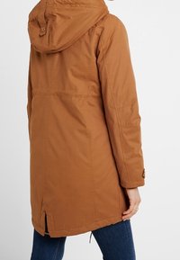 Woman wearing a knee-length brown hooded coat with long sleeves, standing facing away against a plain white background.