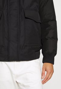 Schwarze gepolsterte Jacke mit zwei aufgesetzten Taschen auf der Vorderseite, gerippten Bündchen und einer glatten Oberfläche. Das Design ist funktional mit einer lässigen Passform.