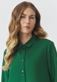 Green collared shirt made of smooth fabric. Features button closures, long sleeves, and a structured collar. No patterns or accents visible.
