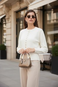 Femme portant des lunettes de soleil et un pull blanc boutonné, tenant un petit sac à main marron dans une rue de la ville avec un arrière-plan flou.
