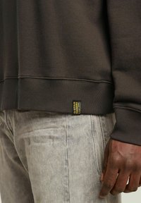 Donkerbruine sweatshirt met ribgebreide manchetten en zoom, voorzien van een klein geel etiket. Gepaard met lichtgrijze jeans, die een gestructureerde stof tonen.
