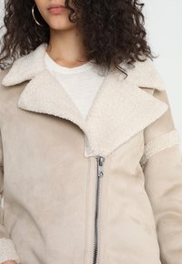 Chaqueta de ante sintético beige con cuello de borreguito, cierre de cremallera y mangas texturizadas. Presenta un diseño estructurado con solapas anchas.