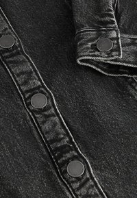 Svart denimtyg med synlig fiskbensstruktur, med svarta metallknappar längs knäppningen och en vikkrage-detalj.