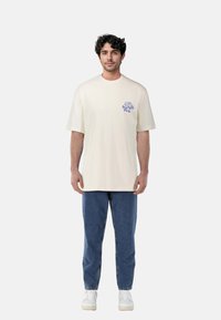 Cream-colored t-shirt with short sleeves and a crew neck, featuring a blue graphic on the left chest. Paired with blue denim jeans.