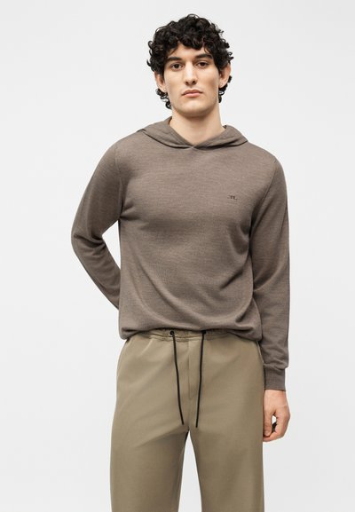 Νέος άνδρας με σγουρά σκουρόχρωμα μαλλιά, φορώντας καφέ φούτερ με κουκούλα και μπεζ παντελόνι, στέκεται μπροστά σε λευκό φόντο.