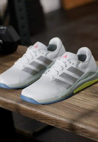 Para białych sneakersów Adidas z szarymi paskami i niebiesko-zielonymi podeszwami spoczywających na drewnianej powierzchni w otoczeniu siłowni.
