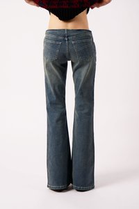 Άτομο που φοράει ξεθωριασμένα μπλε φούστα τζιν, φαίνεται από τη μέση και κάτω, σηκώνοντας ένα σκούρο κόκκινο και μαύρο πουλόβερ, σε απλή λευκή φόντο.