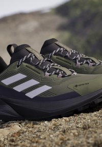 Zapatos de senderismo verdes y negros con una parte superior texturizada, marca de tres rayas en blanco, etiqueta Gore-Tex y una suela robusta para tracción.