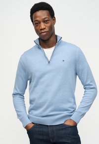 Νεαρός άνδρας που φοράει ανοιχτό γαλάζιο πουλόβερ με φερμουάρ στο λαιμό και σκούρο τζιν, στέκεται με τα χέρια στις τσέπες μπροστά σε λευκό φόντο.