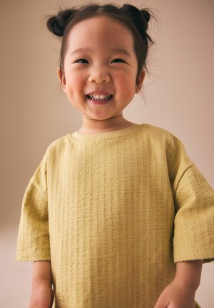 Naeratav noor tüdruk, kellel on juuksed kahes kuklis, kandes tekstuuriga kollast lühikese varrukaga särki tavalise beežika taustaga.