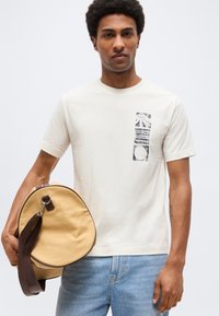 Άνδρας που φοράει λευκό T-shirt με σχέδιο και ανοιχτόχρωμο τζιν κρατάει μπεζ υφασμάτινη τσάντα duffel με καφέ λουράκι, σε λευκό φόντο.