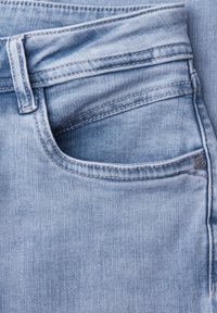 Zbliżenie na jasnoniebieskie jeansy z denimu, pokazujące przednią kieszeń, kieszonkę na monety, szlufkę na pasek oraz metalowe nitowanie.