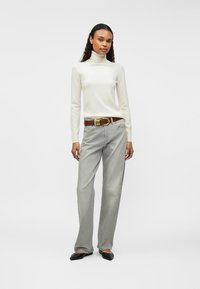 Woman standing wearing a white turtleneck sweater, gray high-waist jeans with a brown belt, and black pointed-toe shoes on a white background.