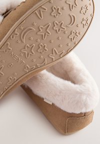 Chaussons en daim brun avec une doublure crème duveteuse et une semelle en caoutchouc texturée ornée de motifs d'étoiles et de lunes. Étiquette de la marque visible.