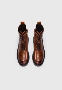 Botas de tobillo de cuero marrón con un acabado suave, punta redonda, cordones en la parte frontal y una suela de goma negra prominente. Incluye cremallera lateral y tira de agarre.