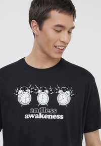 T-shirt in cotone nero con grafica di tre sveglie con effetti sonori, testo "risveglio infinito" in bold bianco.