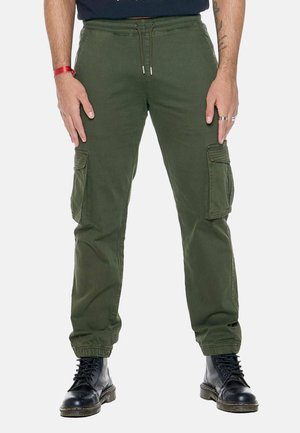 Homme portant un pantalon cargo vert olive avec une taille à cordon et des poignets élastiques, associé à des bottines noires lacées sur un fond uni.