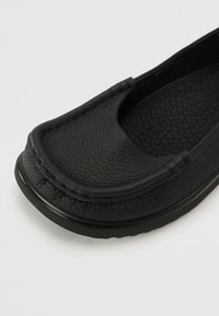 Svart läder slip-on sko med en texturerad yta, rund tå och minimal sömnad längs kanten. Har en flexibel gummisula.