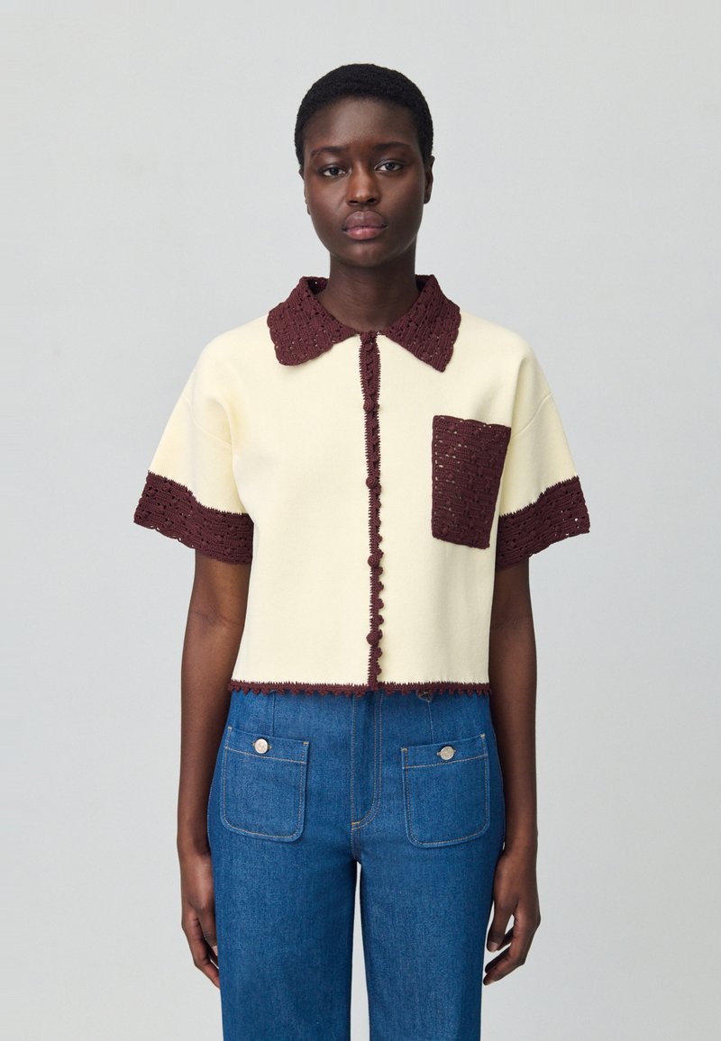 Blusa de manga corta en crema con un collar y bolsillo de textura marrón oscuro, que presenta detalles de botones visibles a lo largo del frente y el dobladillo.