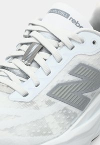 Baskets de course New Balance FuelCell Rebel blanches et grises avec un tissu texturé, une semelle épaisse et des lacets blancs noués en nœud.