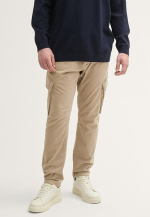 Beige Cargo-Hosen mit zwei Seitentaschen, schmalem Schnitt und elastischen Bündchen, kombiniert mit weißen Sneakers und einem marineblauen Pullover.
