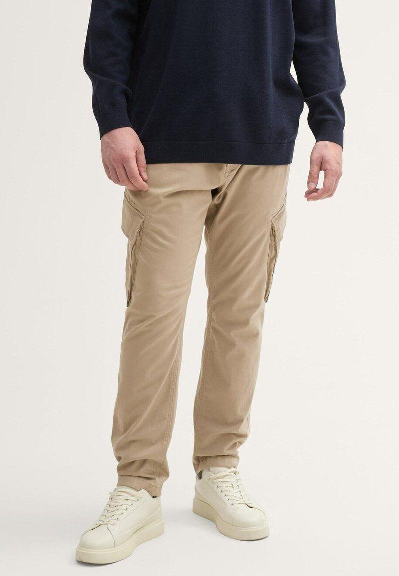 Beige Cargo-Hosen mit zwei Seitentaschen, schmalem Schnitt und elastischen Bündchen, kombiniert mit weißen Sneakers und einem marineblauen Pullover.