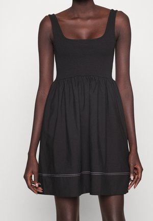 Femme portant une robe noire sans manches avec un décolleté carré et une jupe froncée avec une couture blanche près de l'ourlet.