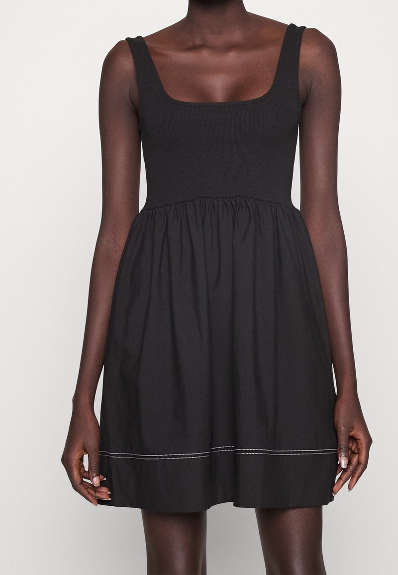 Femme portant une robe noire sans manches avec un décolleté carré et une jupe froncée avec une couture blanche près de l'ourlet.