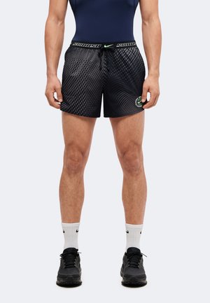 Homme portant un short à carreaux noir et gris avec un logo Nike, des chaussettes blanches Nike, des baskets noires, debout avec les bras détendus sur les côtés.