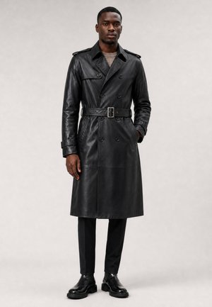 Mann trägt einen langen schwarzen Ledermantel mit Gürtel, schwarze Hose und schwarze Stiefel und steht vor einem schlichten hellen Hintergrund.