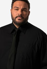 Zwarte overhemd met puntige kraag, lange mouwen en een borstzak. Gecombineerd met een donkergroene stropdas. Smooth textuur.