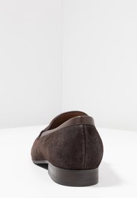 Chaussure en daim marron avec un bout rond, accents en cuir lisse, petit talon et surface texturée, placée sur un fond blanc.