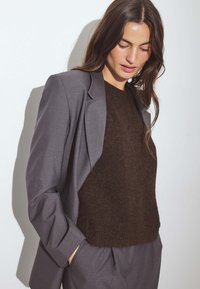 Grauer Blazer mit strukturierten Schultern, kombiniert mit einem dunkelbraunen, strukturierten Pullover. Glatte Stoffe ergänzen den modernen, gestuften Look.