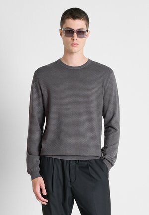 Jonge man met donkere zonnebril, een gestructureerde grijze trui met lange mouwen en losse zwarte broek staat met één hand in de zak.