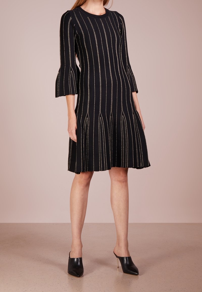 Femme portant une robe noire mi-genou avec des rayures verticales beiges et des manches évasées 3/4, associée à des mules à talons hauts pointues noires.