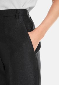 Schwarze Hose mit zartem Glanz, ausgestattet mit einer figurbetonten Taille, Seitentasche und glatter Textur, gezeigt mit einer Hand in der Tasche.