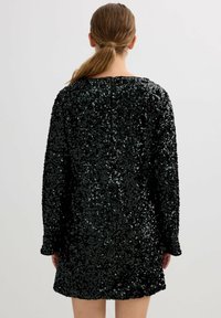 Vestido mini negro con lentejuelas y manga larga, con un corte holgado y diseño de escote redondeado. La superficie texturizada brilla bajo la luz.