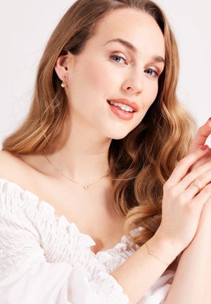 Gouden ketting met een ronde hanger, bijpassende oorbellen en een slanke gouden ring. Het model heeft golvend bruin haar en draagt een witte, gerimpelde top.