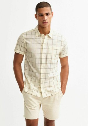Short-sleeve, cream shirt with a grid pattern in brown. Features a button-down collar and a chest pocket. Worn with light beige shorts.