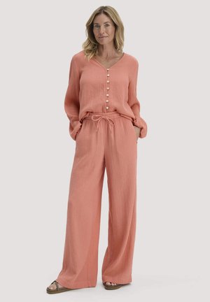 Frau steht und trägt eine lockere, strukturierte, lachsrosa Bluse mit Knöpfen sowie passende, weit geschnittene Hosen mit Kordelzugbund und braune Sandalen.