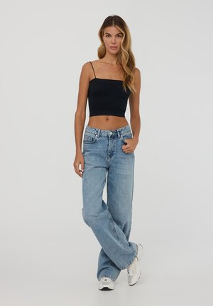 Top corto nero con spalline sottili, abbinato a jeans a gamba larga azzurri e sneakers bianche, su uno sfondo neutro.